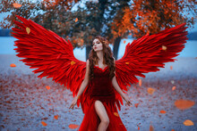 Young Beautiful Woman Fallen Angel Stands In The Park Near A Tree With Orange Falling Leaves. Creative Red Suit, Huge Artificial Bird Wings And Elegant Dress. Wind Magic Whirls The Autumn Foliage