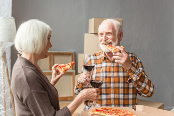 laughing senior couple eating pizza and holding glasses of wine in new house