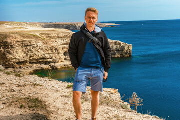 Attractive young man stands on a high cliff overlooking the rocky cliffs arches on the beach and turquoise sea water on the coast