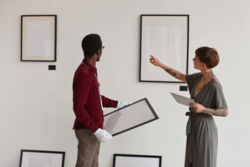 Back view portrait of female art gallery manager instructing worker hanging painting frames on white wall while planning exhibition in museum, copy space