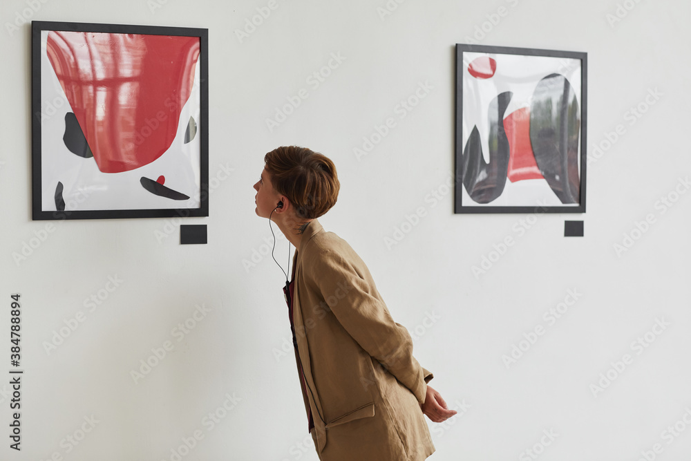 Wide angle portrait of tattooed young woman looking at paintings and listening to audio guide at modern art gallery exhibition, copy space - obrazy, fototapety, plakaty 