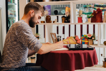 A bearded man sits at a table in a cafe and writes in a notebook.