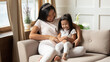 Happy Asian mother and little daughter wearing princess diadems tickling, cuddling, having fun together, sitting on cozy couch at home, family enjoying leisure time, playing funny game