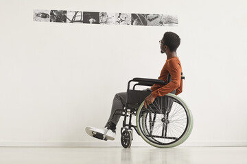 Graphic full length portrait of young African-American man using wheelchair and looking at paintings while exploring modern art gallery exhibition, copy space