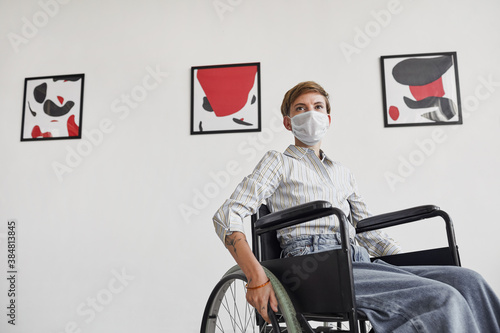 Wide angle portrait of young woman using wheelchair and wearing mask while looking at paintings in modern art gallery