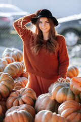 Wall Mural - Portrait of happy woman with ripe orange pumpkin in hands on background of farmers market in brown sweater and hat. Cozy autumn vibes Halloween, Thanksgiving day.