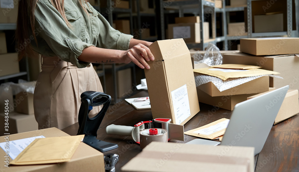 Female small business owner, entrepreneur, shipment delivery dropshipping service worker packing ecommerce order package in shipping cardboard box standing at table in warehouse. Close up view - obrazy, fototapety, plakaty 