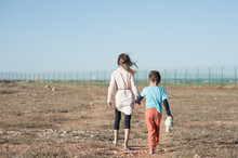 Two Poor Children Family Brother With Toy And Thin Sister Refugee Illegal Immigrant Walking Barefooted Through Hot Desert Towards State Border With Barbed Fence Wire