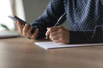 Crop close up of woman sit at desk write make notes study on web internet using smartphone gadget. Smart female handwrite summarize on paper take online course or training on cellphone.