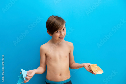 A cheerful boy of 9 years old examines a paper boat. Photo of a cheerful boy who is going to swim on a blue background. Studio photo. boy tourist in swimming trunks