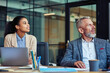 © Kostiantyn - Business people at work. Young mixed race woman and her mature colleague looking away while sitting at desk and working together in the mode office