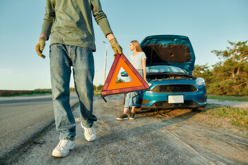 Wall Mural - Cropped shot of man driver putting red warning triangle or emergency stop sign behind his broken car on the side of the road, his girlfriend waiting in the background