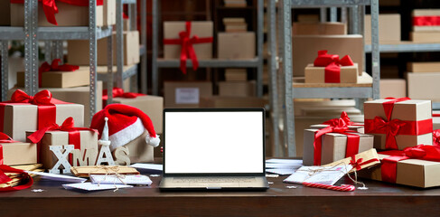 Laptop computer with white blank empty mock up screen on Merry Christmas table with presents gifts boxes in warehouse background. Ecommerce website xmas online shopping and shipping delivery, banner.