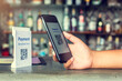 © Koonsiri - Hand holding smartphone to scan Qr code payment tag with blurry soft drink and hand drink at counter bar in coffee shop to accepted generate digital pay without money. Qr code payment concept.