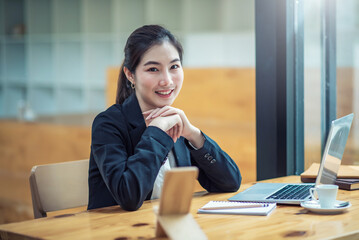 Young pretty asian businesswoman working on laptop while sitting at the table in office. Looking at camera.