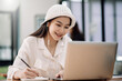© amnaj - Portrait of pretty young asian woman wearing a white hat taking note and using laptop computer at home. Female student studying online at home.