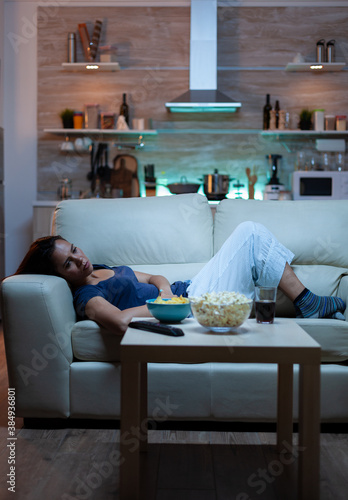 Exhausted woman watching tv show lying on sofa in living room. Tired lonely unhappy young lady in pijamas relaxing on comfortable couch in front of television feeling bored eating snacks late at night