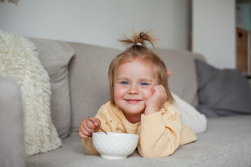 A cute little girl 2 years old in a cozy knitted sweater is having breakfast on the couch. Cozy. Fall.