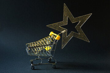 Wall Mural - Shopping cart with a gold transparent star with sequins in the shape of a Christmas tree on a black background. Black Friday, buying gifts for the new year. Space for text