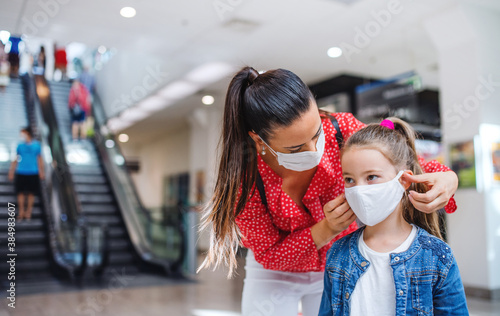 Mother and daughter with face mask standing indoors in shopping center, coronavirus concept.