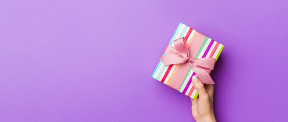 Flat lay of woman hands holding gift wrapped and decorated with bow on purple background with copy space. Christmas and holiday concept