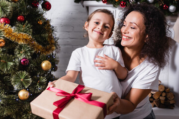 Wall Mural - Little girl with gift box looking at camera near mother and festive pine at home