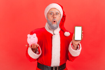 Wall Mural - shocked surprised santa claus holding smartphone with empty screen and piggy bank, cashback, online 