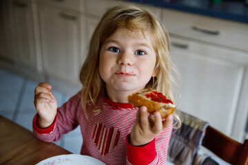 Cute funny toddler girl eats sweet bun for breakfast. Happy child eating bread roll with strawberry jam. Health food for children and kids with selfmade jelly