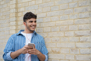 Young handsome asian man using mobile phone, chatting, communication, looking at copy space. Smiling Indian hipster listening music outdoors, standing near wall 