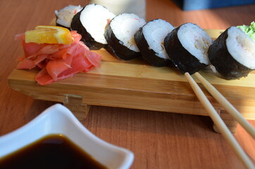 A teenager boy takes sushi roll with Chinese bamboo sticks and drops it into soy sauce, takes a piece of pink ginger. Japanese cuisine on a white porcelain plate next to green wasabi ginger and sauce