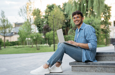 Portrait of happy Indian programmer using laptop computer, internet, working freelance project online, sitting in park. Asian student studying, learning language, using modern technologies outdoors