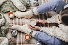 Young Woman Sits On Plaid In Cozy Knitted Woolen White Sweater And Socks Holds Cup Of Cocoa In Her Hands. Hygge New Year, Cozy Christmas, Preparation For Holidays. Candles, Christmas Balls.