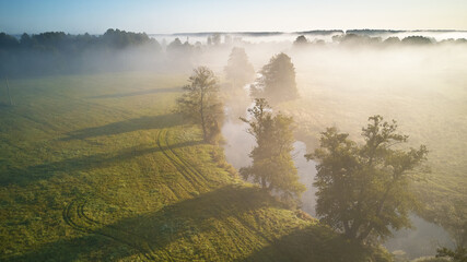  Autumn foggy sunrise aerial view. Small river with trees in meadow and field
