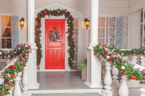Christmas porch decoration idea. House entrance with red door decorated for holidays. Red and green wreath garland of fir tree branches and lights on railing. Christmas eve at home