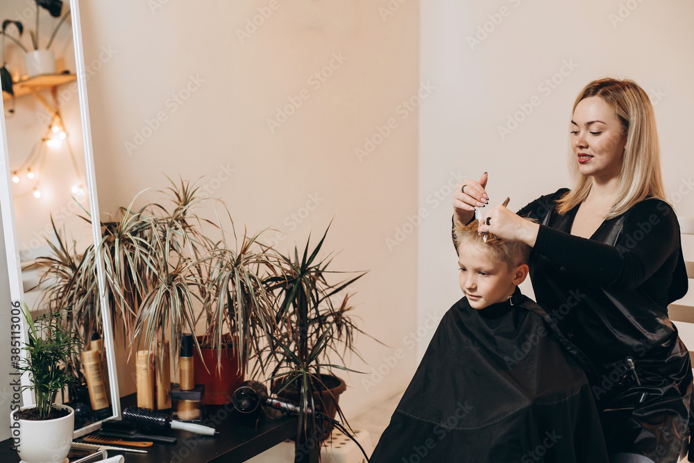 Eight-year-old blonde boy at the barbershop. A hairdresser cuts a haircut at a home beauty salon. Selective focus. - obrazy, fototapety, plakaty 