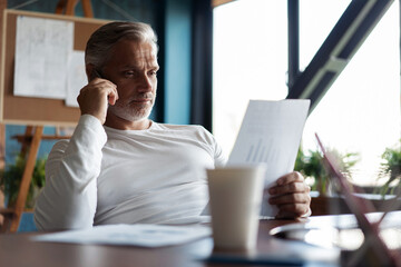 Canvas Print - Grey-haired mature businessman sitting at table, working on computer, talking on smartphone. Telephone conversations