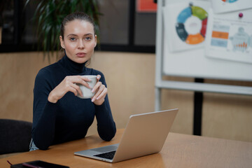 working on laptop computer. holding coffee in hands thinks over new statistics plan site. serious beautiful young intelligent woman of Caucasian appearance ethnicity, sitting alone in the office.