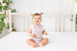 © Any Grant - cheerful baby girl six months old sitting in a bright beautiful room on a white bed in a lace bodysuit and smiling