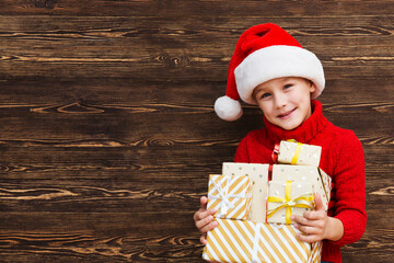 Kid boy  in a Santa hat  with Christmas gift boxes on a wooden background. Christmas shopping.  Copy space. new normal.