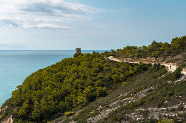 Wall Mural - Landscape of Sierra de Irta Natural Park full of green trees with the Badum sentinel tower in the background on a summer day with blue sky, Castellon, Spain