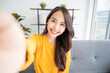 © Monster Ztudio - Pretty young asian female with big smile sitting at living room. She having fun taking light cheerful selfie on blurred background