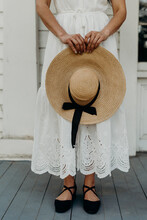 Petite Woman Wearing A White Lace Dress And Black Shoes Holding A Straw Hat Standing On An Old Porch