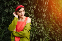 Attractive Young Fashion Style Teenager Girl Shyly Embarrassed Looking Down. Red And Green. Puberty Problems. Feminine Slim Figure Body. Piercing In The Nose And A Mole On The Face. 
Nature Background