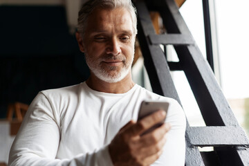 Wall Mural - Casual Grey-haired Mature businessman using smartphone standing by the window