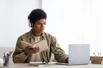 Focused teenage student in headset reading textbook during online lesson on laptop at home, blank space
