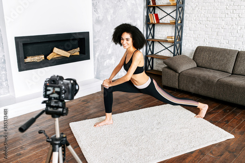 A cheerful African-American woman records fitness video classes on camera. A mixed race girl in sportswear is doing stretching exercises. Sports vlogging concept, yoga coach online