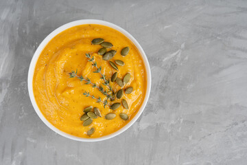 Wall Mural - Vegetarian homemade organic pumpkin soup of orange color made of pureed vegetables decorated with seeds and fresh thyme served in bowl on concrete background. Image with copy space, horizontal