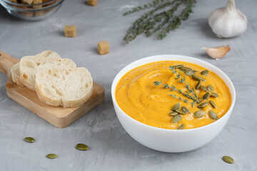 Wall Mural - Creamy organic pumpkin soup made of pureed vegetables decorated with seeds and fresh thyme served in white bowl with bread slices, croutons and garlic on concrete background. Horizontal orientation