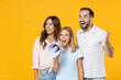 © ViDi Studio - Surprised excited young parents mom dad with child kid daughter teen girl in basic t-shirts scream in megaphone spreading hands isolated on yellow background. Family day parenthood childhood concept.