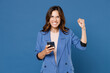 Happy young brunette woman 20s wearing basic jacket standing using mobile cell phone typing sms message doing winner gesture looking camera isolated on bright blue colour background, studio portrait.